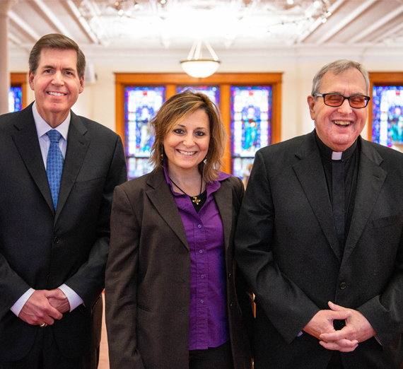 Группа из пяти человек стоит вместе в ярко освещенном коридоре больницы, улыбаясь в камеру. Двое носят стетоскопы и медицинскую одежду, один — толстовку Университета Гонзага, остальные одеты в повседневную или деловую одежду, представляя смесь студентов и медицинских работников.